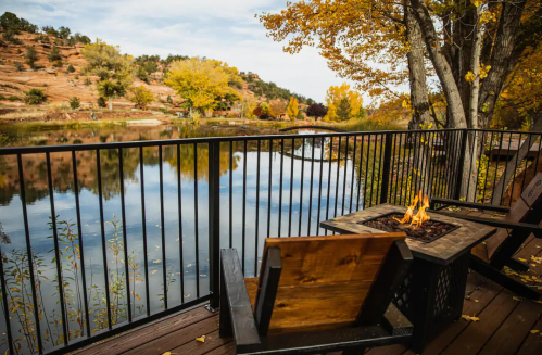 A cozy outdoor seating area by a calm lake, featuring a fire pit and autumn foliage reflecting on the water.